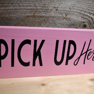 Pode incluir: Uma placa de madeira rosa com letras pretas que diz "PICK UP Here".