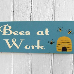 Può includere: Un cartello di legno blu con il testo "Bees at Work" dipinto in bianco. Il cartello presenta anche un alveare giallo e nero e tre api gialle e nere.