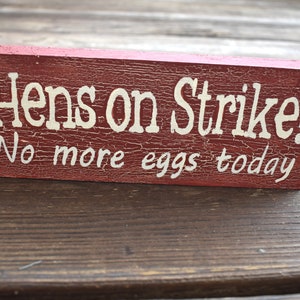 May include: A red wooden sign with white lettering that reads "Hens on Strike! No more eggs today".