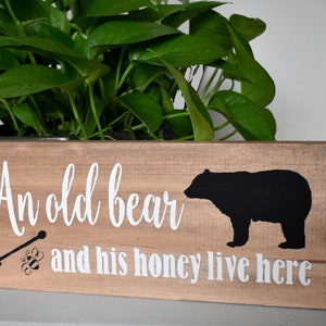 May include: A wooden sign with the text "An old bear and his honey live here" in white paint. A black silhouette of a bear is to the right of the text. A small black and white illustration of a honey dipper and a bee is to the left of the text.
