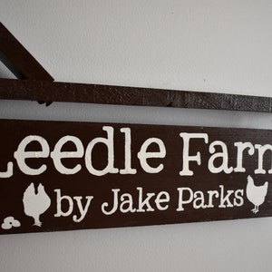 Puede incluir: Un letrero de madera marr&oacute;n con letras blancas que dice "Leedle Farm by Jake Parks". El letrero tiene dos siluetas de pollos blancas y dos huevos blancos en &eacute;l.