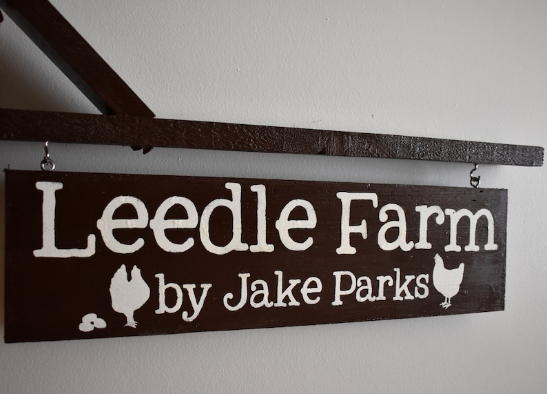 Puede incluir: Un letrero de madera marr&oacute;n con letras blancas que dice "Leedle Farm by Jake Parks". El letrero tiene dos siluetas de pollos blancas y dos huevos blancos en &eacute;l.