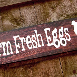 May include: A rustic wooden sign with the text "Farm Fresh Eggs" in white paint. A white silhouette of a rooster is painted on the right side of the sign.