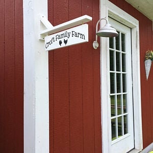 Puede incluir: Un letrero de madera blanco con el texto "Overt Family Farm" y dos siluetas de pollos negras. El letrero est&aacute; sujeto a un poste blanco y montado en un edificio rojo.