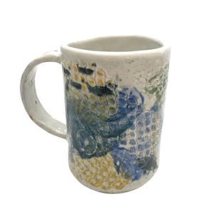 May include: A handmade ceramic mug with a white base and a curved handle. The mug features a textured surface with blue, green, and yellow abstract patterns. The mug is designed for hot beverages.