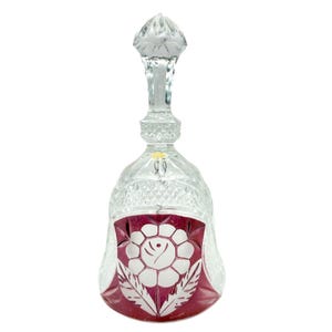 May include: A clear and cranberry red cut-to-clear crystal bell featuring a faceted handle and a diamond-cut pattern on the upper body. The lower section displays a prominent white floral motif with leaves etched into the deep red glass surface.