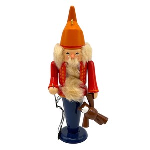 May include: A wooden nutcracker figurine featuring a tall orange pointed hat, a red jacket with yellow dot accents, and a dark blue base. The figure has a fluffy beige beard and hair, and holds a small brown bear toy in its left hand.