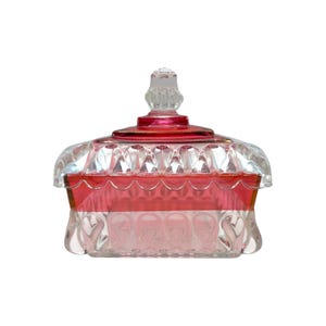 May include: A rectangular glass candy dish featuring a clear base and lid with a vibrant red band around the center. The lid is topped with a clear, faceted finial, and the entire piece is decorated with a repeating embossed scalloped pattern.