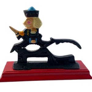 May include: A vintage nutcracker in the shape of a soldier, painted in black, gold, blue, and flesh tones. The nutcracker is mounted on a red wooden base. The soldier figure has a blue hat and a blonde wig.