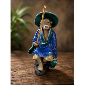 May include: A ceramic figurine of an elderly person with a white mustache wearing a wide-brimmed dark green hat and a blue robe. The figure is seated on a dark rock, holding a thin, light-colored bamboo fishing pole in one hand.