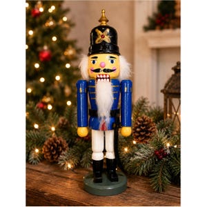 May include: A wooden nutcracker figurine wearing a black helmet with a gold finial and red gem detail. The figure has a blue jacket with gold trim, white trousers, black boots, and a long white beard. It stands on a circular green base.