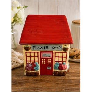 May include: A ceramic cookie jar shaped like a flower shop with a bright red roof. The cream-colored facade features the text "FLOWER SHOP" above a red door labeled "NO 3 ROSENIP LANE" with an "OPEN" sign, flanked by windows and flower boxes.