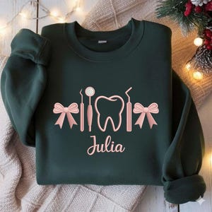 May include: Dark green sweatshirt with a dental theme. The design includes a tooth, dental tools, and bows in a light pink color. The name "Julia" is written in cursive below the design.