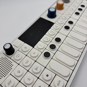 May include: A white and silver electronic music synthesizer with various colored knobs and buttons. The device has a grid of white keys with black symbols and a black screen. The knobs are in red, gray, orange, and blue.