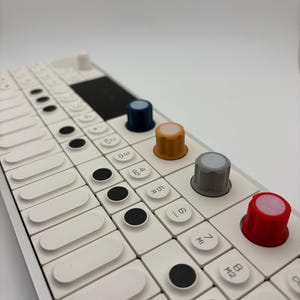Puede incluir: Primer plano de un sintetizador de música electrónica blanco con una cuadrícula de teclas blancas y botones circulares negros. Cuenta con varias perillas de colores: azul, beige, gris y rojo. El dispositivo está diseñado para la producción y la interpretación musical.