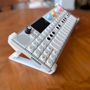 Op de afbeelding: Een witte elektronische synthesizer met een rechthoekige interface, voorzien van een zwarte knop, een klein scherm en meerdere witte toetsen en knoppen. Het apparaat staat op een doorzichtige witte kunststof standaard in een schuine stand.