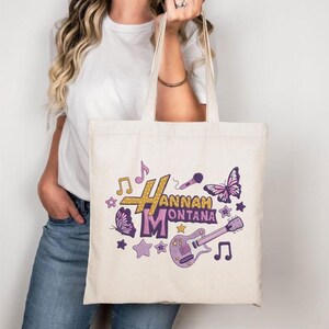May include: Cream-colored tote bag featuring the text "Hannah Montana" in gold and purple, with musical notes, stars, a microphone, a butterfly, and a guitar graphic. The bag has two handles and is held by a person.