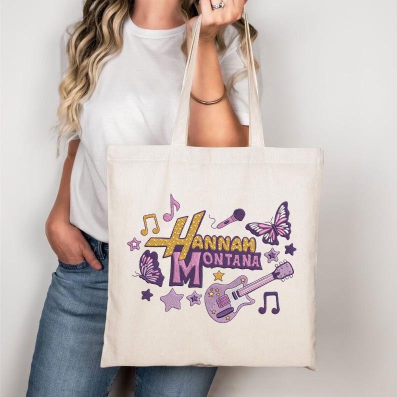 May include: Cream-colored tote bag featuring the text "Hannah Montana" in gold and purple, with musical notes, stars, a microphone, a butterfly, and a guitar graphic. The bag has two handles and is held by a person.