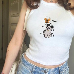 May include: A white sleeveless tank top featuring a graphic print of a ghost with Mickey Mouse ears holding a pumpkin balloon. The design includes small black bats and star shapes surrounding the central ghost figure on the front of the shirt.