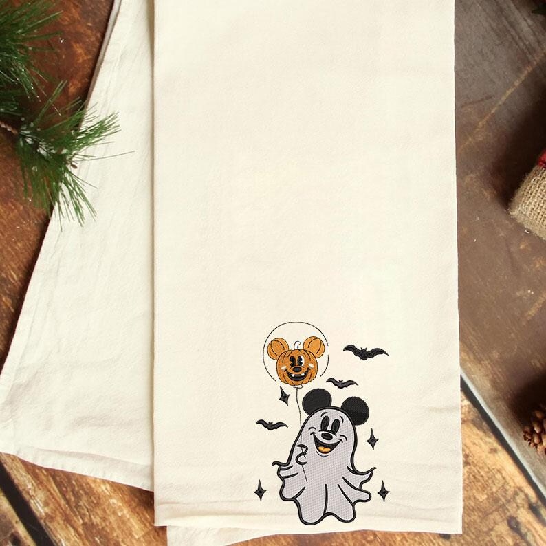 May include: A cream-colored kitchen towel featuring an embroidered design of a ghost wearing black mouse ears and holding a pumpkin balloon with mouse ears. The design includes small black bats and four-pointed stars surrounding the figures.