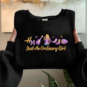 May include: Black sweatshirt with the text "Hm Just An Ordinary Girl" in gold and a graphic of a woman with a microphone, guitar, boots, and a hat. The graphic is in purple, gold, and white.