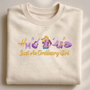 May include: Cream-colored sweatshirt with embroidered design. Features a guitar, microphone, boots, and a hat, with the text "Just An Ordinary Girl" in gold and purple. The design includes musical notes and a stylized female figure.