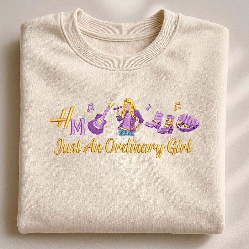 May include: Cream-colored sweatshirt with embroidered design. Features a guitar, microphone, boots, and a hat, with the text "Just An Ordinary Girl" in gold and purple. The design includes musical notes and a stylized female figure.