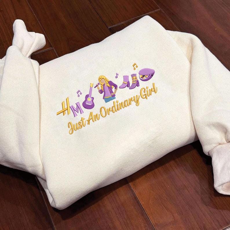 May include: Cream-colored sweatshirt with embroidered design. The design features a musical theme with a guitar, microphone, boots, and a hat, along with the text "HM Just An Ordinary Girl" in gold and purple.
