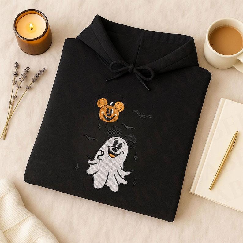 May include: A black hooded sweatshirt featuring an embroidered design of a white ghost character with mouse ears and a smiling orange pumpkin with mouse ears. The design includes small embroidered bats and stars surrounding the characters.