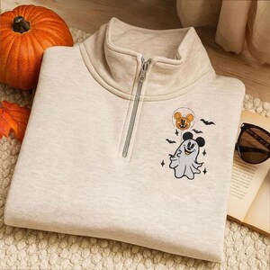 May include: A beige quarter-zip sweatshirt featuring a graphic of a ghost wearing Mickey Mouse ears and holding a pumpkin balloon, surrounded by small bats and stars. The sweatshirt has a high collar with a silver-toned metal zipper closure.