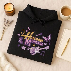 May include: Black hoodie with a colorful embroidered design featuring the name "Hannah Montana", musical notes, stars, butterflies, and a guitar. The design incorporates gold, purple, and white colors.