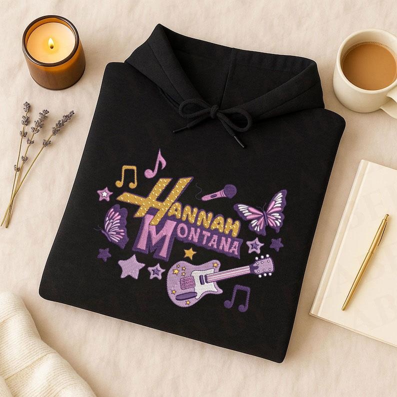 May include: Black hoodie with a colorful embroidered design featuring the name "Hannah Montana", musical notes, stars, butterflies, and a guitar. The design incorporates gold, purple, and white colors.