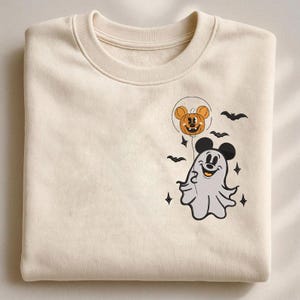 May include: A cream-colored crewneck sweatshirt featuring an embroidered design on the chest. The embroidery depicts a white ghost wearing black mouse ears, holding a string attached to an orange pumpkin balloon with mouse ears, surrounded by bats.