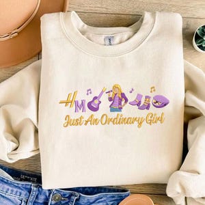 May include: Cream-colored sweatshirt with a graphic design featuring a woman with a microphone, musical notes, a guitar, boots, and a hat. The text "HM Just An Ordinary Girl" is embroidered in gold and purple.