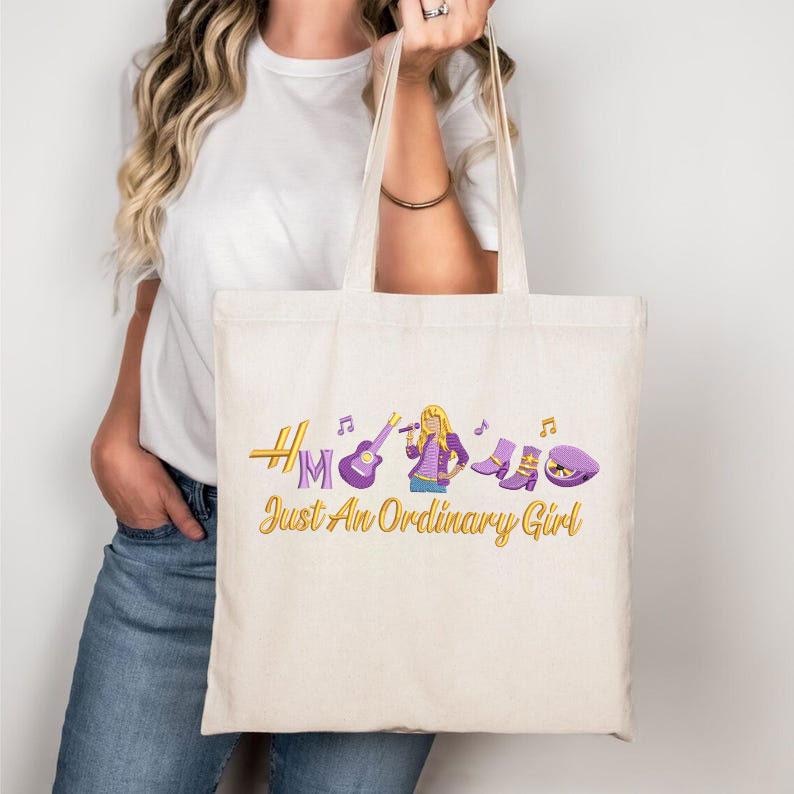 May include: Cream-colored tote bag with a design featuring a singer, musical notes, a guitar, and boots. The text "Just An Ordinary Girl" is in gold script. The bag is held by a person wearing a white t-shirt and blue jeans.