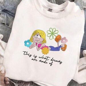 K&ouml;nnte beinhalten: Wei&szlig;es Sweatshirt mit einer Cartoon-Illustration eines blonden M&auml;dchens, das von Blumen umgeben liegt. Das M&auml;dchen tr&auml;gt ein rosa Oberteil, blaue Hosen und orangefarbene Schuhe. Der Text "this is what dreams are made of" steht darunter.