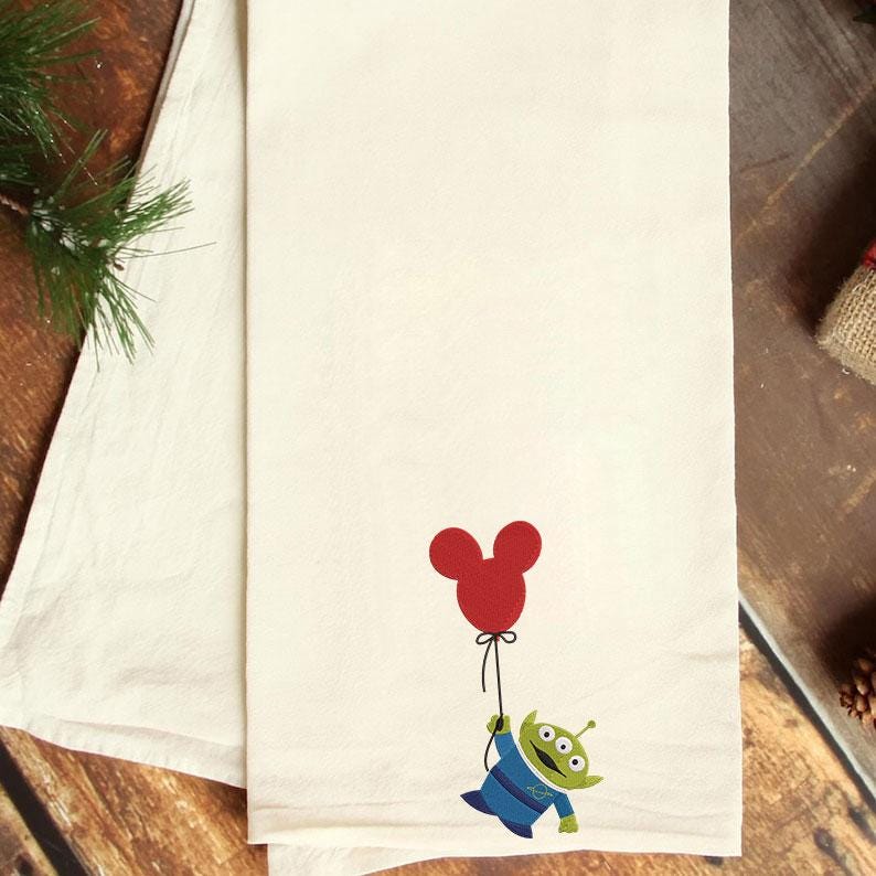 May include: Cream-coloured kitchen towel featuring a cartoon alien holding a red Mickey Mouse balloon. The alien is green with three eyes and wears a blue uniform. The balloon is red. The towel is folded.