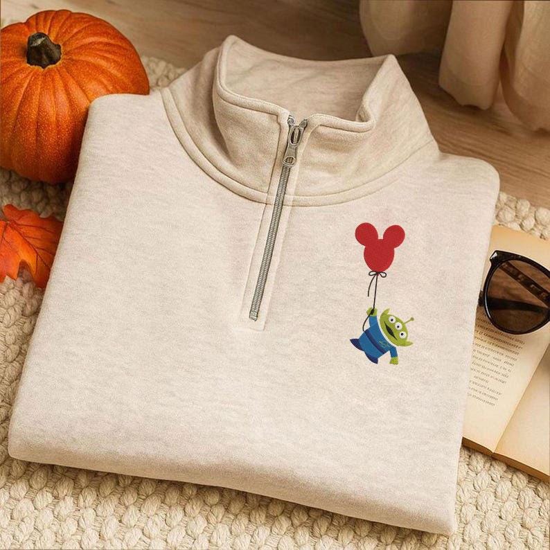 May include: A beige quarter-zip sweatshirt with a cartoon alien holding a red Mickey Mouse balloon. The sweatshirt has a high collar and a silver zip. A pumpkin and autumn leaves are in the background.