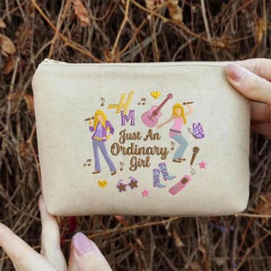 Puede incluir: Bolsa beige con cremallera y tem&aacute;tica musical. El dise&ntilde;o incluye ilustraciones de una guitarra, un micr&oacute;fono y una mariposa, junto con el texto "Just An Ordinary Girl". La bolsa es sostenida por una persona.