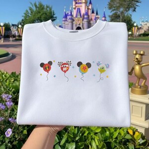 May include: White sweatshirt featuring four Mickey Mouse-themed balloon designs. Each balloon has a different design, including a red balloon with a black Mickey head, a checkered balloon with the number "95", and two other balloons with unique patterns. The sweatshirt is held up by a hand.