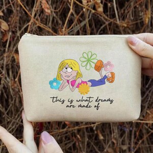 K&ouml;nnte beinhalten: Beige Canvas-Beutel mit Rei&szlig;verschluss. Der Beutel zeigt eine Cartoon-Illustration eines blonden M&auml;dchens, das liegt und von Blumen umgeben ist. Der Text "This is what dreams are made of" ist darunter gedruckt.