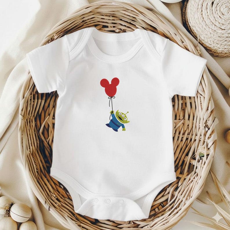 May include: White baby bodysuit with a cartoon alien character holding a red balloon. The alien is green with three eyes and is wearing a blue outfit. The bodysuit is displayed in a woven basket.