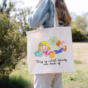 K&ouml;nnte beinhalten: Cremefarbene Tragetasche mit einer Stickerei eines Cartoon-Charakters mit blonden Haaren, einem rosa Oberteil und blauen Hosen, umgeben von bunten Blumen. Der Text "this is what dreams are made of" steht darunter.