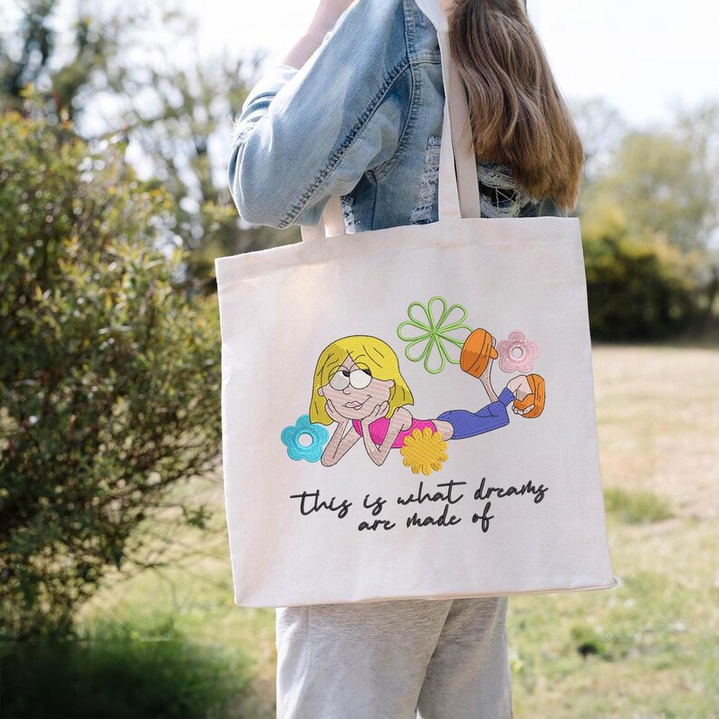 K&ouml;nnte beinhalten: Cremefarbene Tragetasche mit einer Stickerei eines Cartoon-Charakters mit blonden Haaren, einem rosa Oberteil und blauen Hosen, umgeben von bunten Blumen. Der Text "this is what dreams are made of" steht darunter.