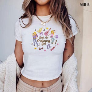 Puede incluir: Top corto blanco con un gr&aacute;fico colorido que dice "Just An Ordinary Girl". El dise&ntilde;o presenta dos figuras de dibujos animados con instrumentos musicales, notas musicales y otros elementos decorativos. La camiseta es de manga corta.