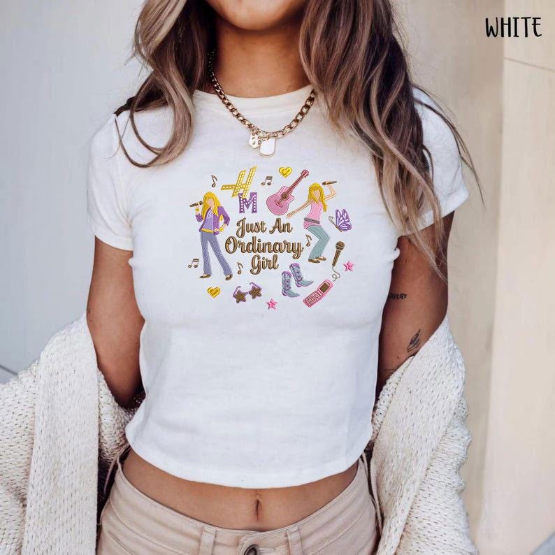 Puede incluir: Top corto blanco con un gr&aacute;fico colorido que dice "Just An Ordinary Girl". El dise&ntilde;o presenta dos figuras de dibujos animados con instrumentos musicales, notas musicales y otros elementos decorativos. La camiseta es de manga corta.
