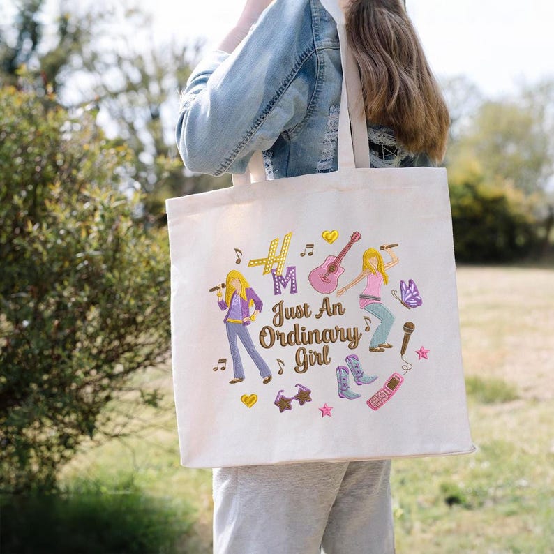 Puede incluir: Un bolso tote de color crema con un gr&aacute;fico colorido. El dise&ntilde;o incluye el texto "Just An Ordinary Girl" e ilustraciones de una chica con un micr&oacute;fono, una guitarra, notas musicales y otros elementos decorativos.