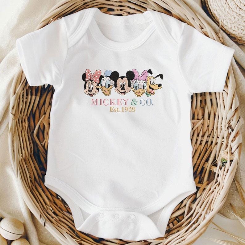 May include: White baby bodysuit featuring a colorful graphic of Mickey Mouse, Minnie Mouse, Donald Duck, Daisy Duck, and Goofy. The text "Mickey & Co. Est. 1928" is printed below the characters. The bodysuit is short-sleeved and has a snap closure.