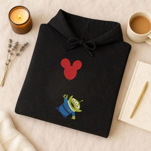 May include: Black hooded sweatshirt with a red Mickey Mouse balloon and a green alien character. The sweatshirt has a drawstring hood. The design is embroidered on the front.