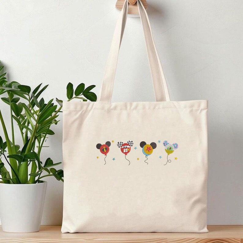 May include: Cream-colored tote bag with four colorful balloon designs. Each balloon features a Mickey Mouse head and various designs, including a checkered flag and the number 95. The bag has long handles.